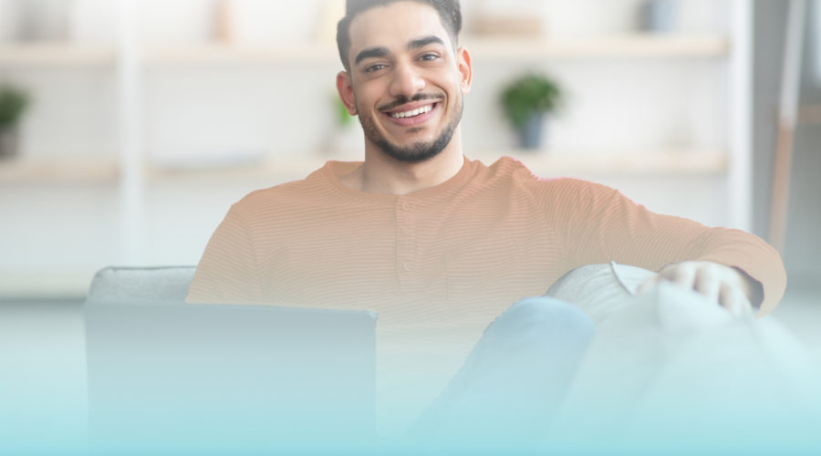 Smiling arab guy reclining on sofa with laptop at home, copy space. Handsome young middle-eastern man enjoying weekend, using notebook, watching moovie or surfing on dating website